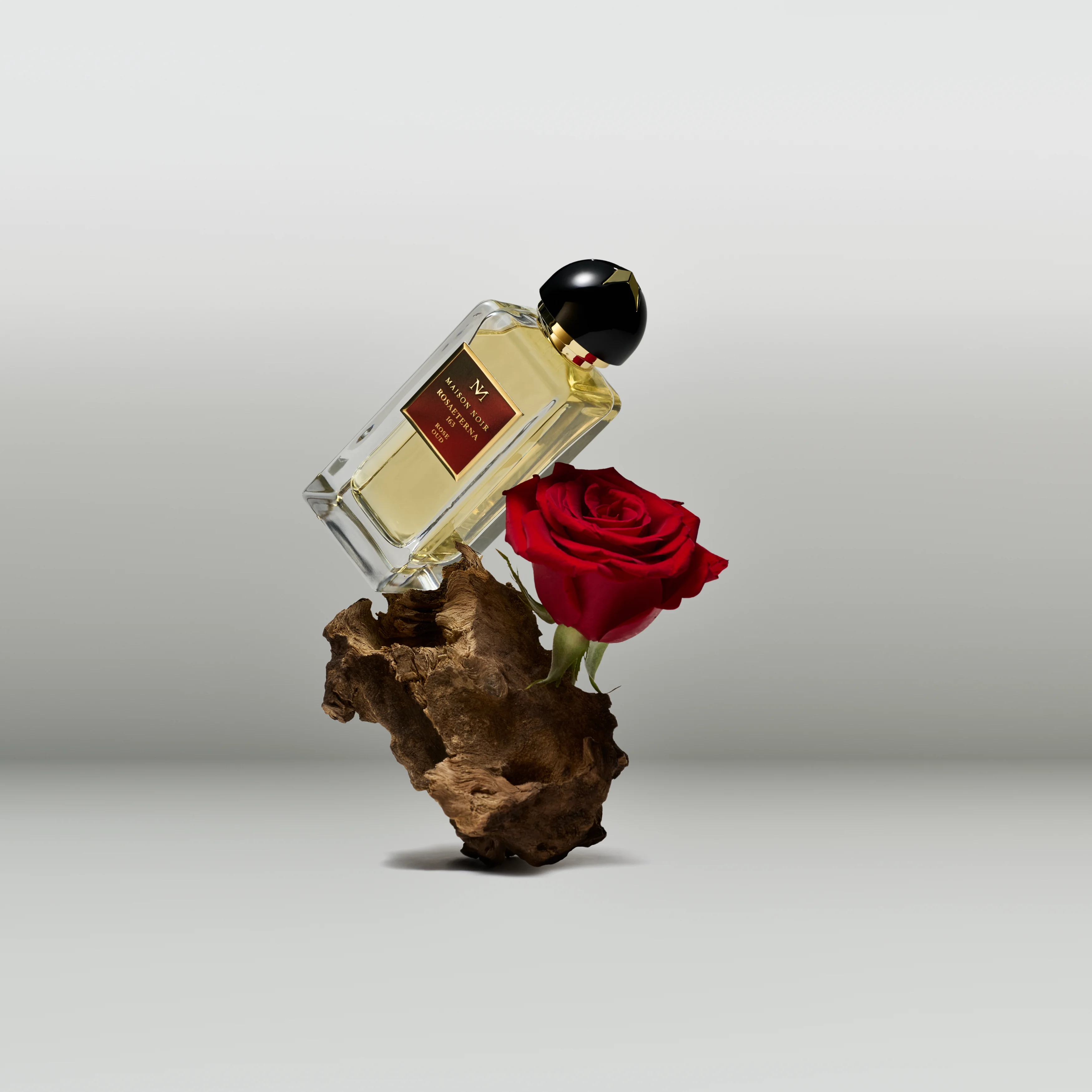 A flacon balanced on weathered wood beside a deep red rose, a floral note meeting a woody base